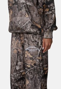 Camo hoodie en joggingbroek in bruin, grijs en zwart, met een textuurstof en een wit logo op de broek.