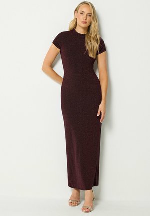 Donna con capelli biondi lunghi, che indossa un vestito lungo aderente a maniche corte color viola scuro glitterato e tacchi beige aperti, in piedi contro un muro bianco.