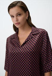 Korte mouw shirt met een kraag, gemaakt van lichte stof met een geometrisch patroon in rood en marineblauw. Soepele textuur.