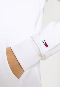 Sudadera blanca con puños acanalados; presenta un pequeño logotipo bordado en rojo, blanco y azul. Textura suave, diseño unisex.