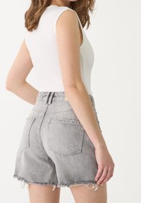 Femme portant un haut sans manches blanc et un short en denim gris taille haute avec des bords effilochés, vue de mi-dos à mi-cuisse.