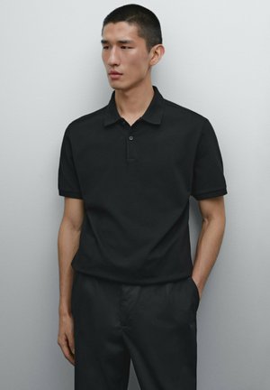 Polo nero a maniche corte, con una chiusura a due bottoni e un tessuto morbido e textured. Indossato con pantaloni scuri abbinati.