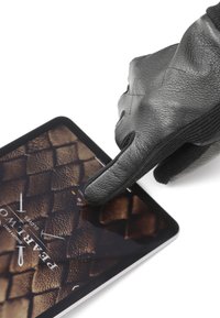 Guanto di pelle nera che tocca lo schermo di un tablet che mostra uno sfondo marrone texturizzato con un motivo che ricorda un materiale intrecciato.