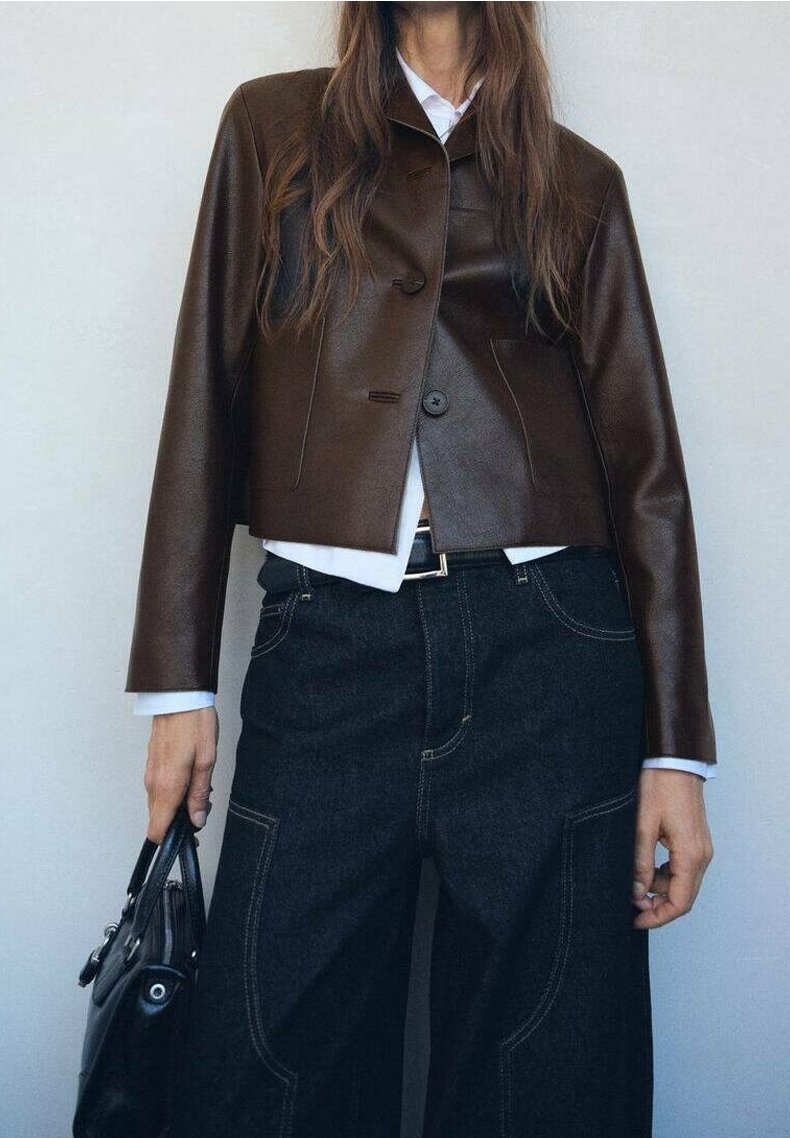Personne portant une veste en cuir marron foncé sur une chemise blanche, avec un pantalon noir taille haute à larges jambes, tenant un sac à main noir dans la main gauche.