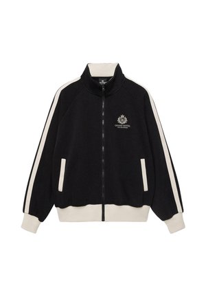 Zwarte jas met rits, witte strepen op de mouwen, witte manchetten en zoom, en het logo "Grand Hotel Les Bains" op de borst.