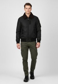 Giubbotto bomber nero con colletto in pelliccia marrone, chiusura con zip e tasche laterali, abbinato a pantaloni cargo verde oliva e stivali neri.