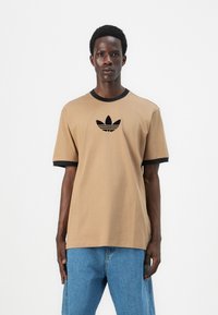 Rudas trumpas rankovės marškinėliai su juodu įsiūtu apykaklės ir rankovių klokeliu. Centrinėje krūtinės dalyje yra juodas adidas logotipas.