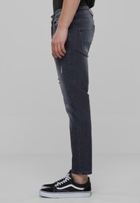 Sötét mosású farmernadrág enyhe kopásokkal, slim fit, öt zsebes kivitelben. Fekete, lapos talpú sportcipővel párosítva, fehér díszítésekkel.