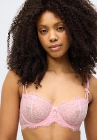 Ροζ δαντελένιο σουτιέν με λουλουδάτα μοτίβα, διαφανείς κύπελλοι, λεπτή κυματιστή διακοσμητική κορδέλα και ρυθμιζόμενους ιμάντες. Μαλακή υφή με φιόγκο στο κέντρο.