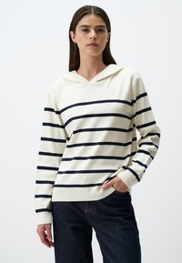 Gestreifter Kapuzenpullover in Creme mit marineblauen Streifen, mit V-Ausschnitt, langen Ärmeln und lockerer Passform, kombiniert mit dunkelblauen Jeans.