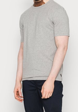 Homme portant un t-shirt uni gris clair à manches courtes et un pantalon bleu marine foncé, debout devant un fond neutre.