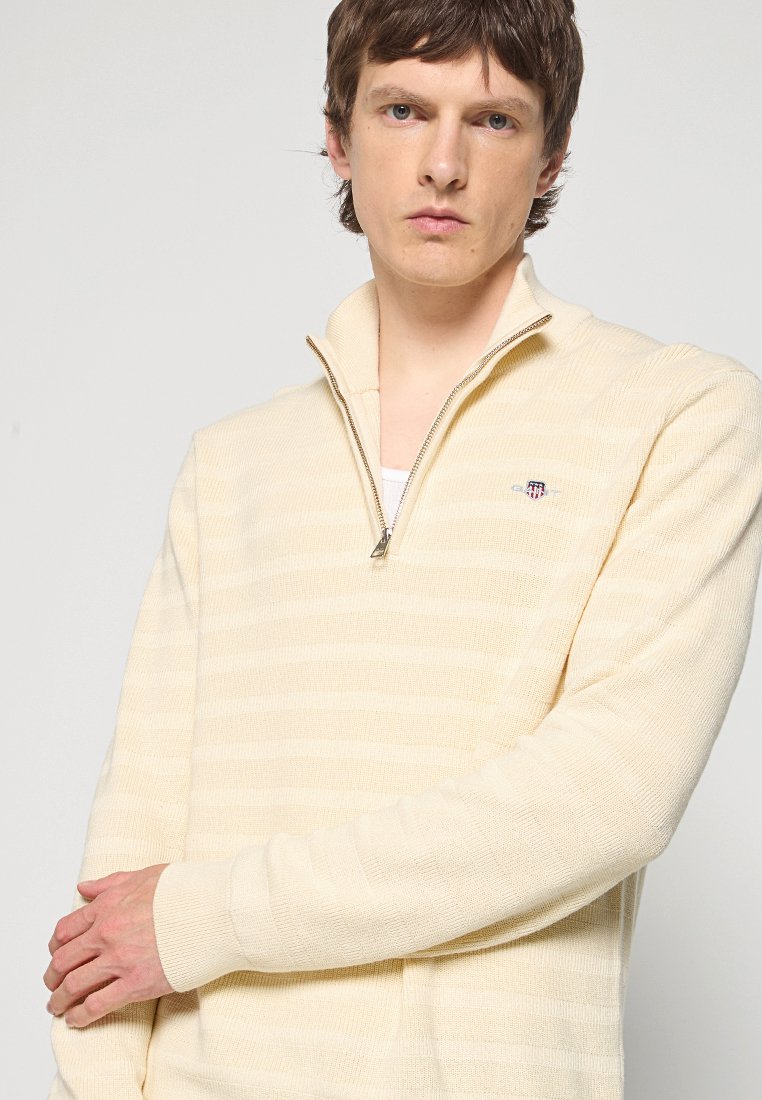 Jeune homme avec des cheveux bruns courts portant un pull à zip quart beige clair avec de fines rayures horizontales et un petit emblème sur la poitrine.