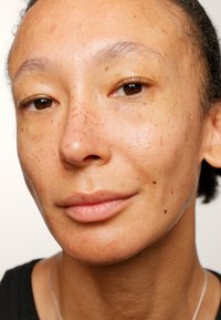 Nærbilde av en persons ansikt med jevn hudtone, subtile fregner, mandelformede brune øyne og glatte lepper. Lys reflekteres fra huden.