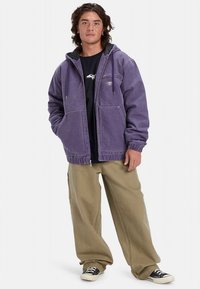 Vijolična jopa s kapuco iz mehke tkanine, z dvema sprednjima žepoma in ohlapnim krojem, združena s širokimi bež hlačami in črnimi supergami.