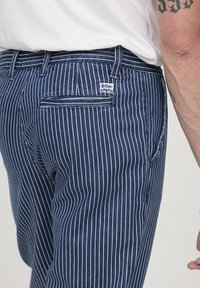Homme portant un jean à rayures verticales bleu et blanc avec une étiquette Lee Cooper sur la poche arrière, bras tatoué visible.