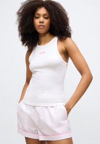 Λευκό αμάνικο μπλουζάκι με κόκκινο λογότυπο "Calvin Klein" και ροζ βερμούδα με ροζ τρέσα. Και τα δύο είναι φτιαγμένα από μαλακό, ελαφρύ ύφασμα.