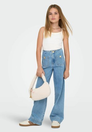 Ung jente med langt blond hår iført hvit singlet, høytlivs vide jeans, hvite joggesko, holder en hvit fluffy håndveske.