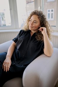 Femme aux cheveux bouclés portant un t-shirt noir, assise confortablement dans un fauteuil clair près d'une fenêtre, regardant sur le côté et touchant son oreille.