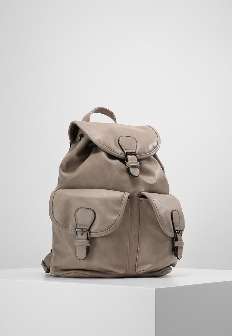 Sac à dos en cuir beige avec trois poches à sangles boucles, une poignée supérieure et des bretelles réglables sur un piédestal blanc sur fond gris.