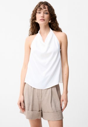 Kvinde med langt, krøllet hår bærer en hvid halterneck-top og beige plisserede shorts, stående mod en ensfarvet lys baggrund.
