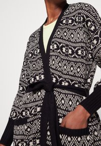 Pletený kardigan v černé a bílé barvě s propracovanými vzory, otevřený přední díl, zavazovací pásek a kapsy. Měkká textura a dlouhé rukávy.