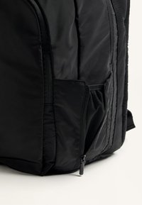 Fekete hátizsák, párnázott külsővel, cipzáras zsebbel, hálós díszítéssel, valamint egy elegáns dizájnnal, tartós szövetből és varrási részletekkel.