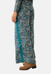 Pantalones de pierna ancha con estampado floral en verde azulado, con una franja lateral de color verde azulado en contraste y una tela suave y texturizada. Combinados con botas de tobillo marrones.