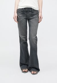 Άτομο που φοράει ξεθωριασμένο μαύρο τζιν bootcut με μπροστινές τσέπες και ασημένια σανδάλια, στέκεται μπροστά σε απλό λευκό φόντο.