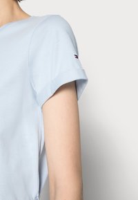 Jasnoniebieska koszula z krótkim rękawem, z podwiniętym mankietem i małym logo na górnej części ramienia, noszona przez osobę na jednolitym tle.