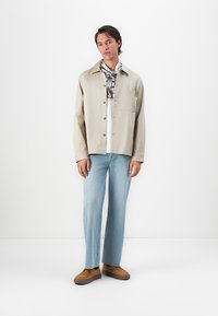 Man stands against white background wearing beige button-up jacket, graphic shirt, light blue jeans, tan slip-on shoes, and black-and-white scarf.