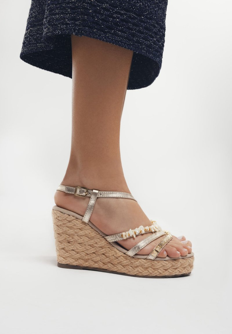 Sandales à talon compensé avec une semelle en jute texturé, présentant des brides métalliques et des accents décoratifs. Les couleurs comprennent l'or et le beige. Design à bout ouvert.
