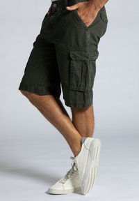 Shorts cargo en coton vert foncé avec une coupe décontractée, poches latérales et un détail de bouton. Assorties avec des baskets blanches.