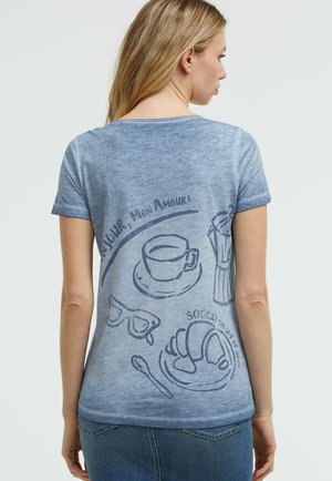 Woman with long blonde hair wears a blue T-shirt with dark blue prints of a coffee cup, croissant, sunglasses, and French text.