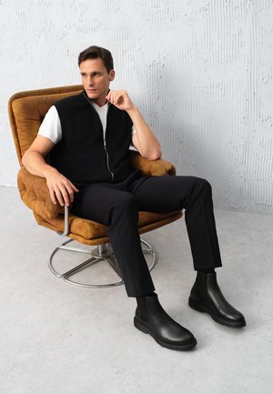 Chaleco negro acanalado con cremallera sobre una camiseta blanca, combinado con pantalones negros y elegantes botas de tobillo negras, sentado en una silla giratoria marrón.