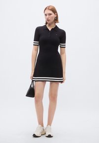 Melnas adītas polokleita ar īsām piedurknēm, baltām svītrām uz piedurknēm un apakšmalā, pieguļošs dizains, kombinēta ar krēmkrāsas sporta apaviem un melnu somu.