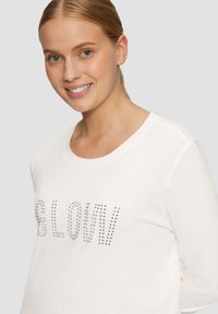 Witte langemouwtop met de tekst "BLOW" in zilveren studs. Zacht katoenen materiaal met een ronde halslijn en een relaxte pasvorm.