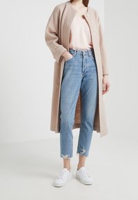 Dlouhý béžový kabát s velkými knoflíky, vrstvený přes světle růžový top, v kombinaci s modrými džíny s vysokým pasem a rozpáranými lemy, a bílými teniskami.