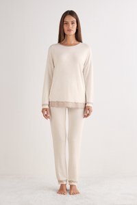 Ljust beige lounge-set som innehåller en långärmad topp med spetsdetaljer vid fållen och matchande byxor med spetsaccenter vid ärmsluten. Mjuk textur.