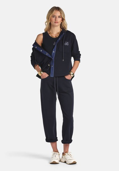 Woman wearing a navy blue sleeveless top, matching jacket partially off one shoulder, drawstring pants, gold jewelry, and white sneakers.