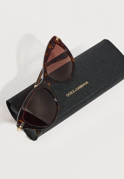 Γυαλιά ηλίου σε σχήμα cat-eye με καραμελί μαρμάρινα πλαίσια και σκούρους καφέ φακούς, τοποθετημένα πάνω σε μαύρη υφασμάτινη θήκη Dolce & Gabbana με χρυσά γράμματα.