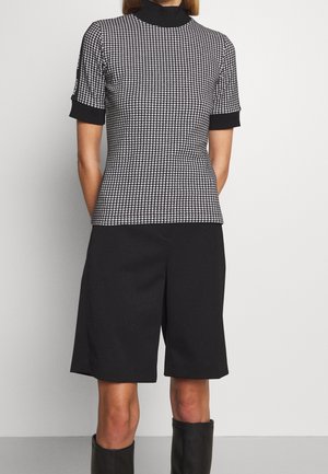 Person wearing a black and white houndstooth short-sleeve top with black collar, black knee-length shorts, and black boots against a plain background.