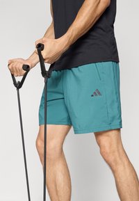 Moški z oblekami v teal Adidas kratkih hlačah in črni majici, ki uporablja črne elastične trakove za vadbo.