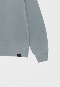 Sudadera azul clara con cuello redondo, puños y bajo acanalados. Presenta una pequeña etiqueta de marca negra en el lado inferior. Textura suave.