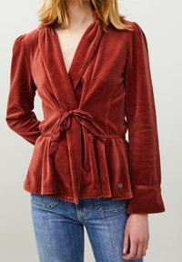 Top de terciopelo rojo con un cierre atado en la cintura, detalles de peplum y mangas largas. Combinado con jeans de denim azul.