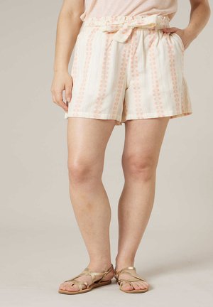 Vrouw in crème korte broek met roze verticale strepen en een geknoopt ceintuur, gecombineerd met beige platte sandalen, staand tegen een neutrale achtergrond.