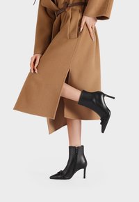 Stivali alla caviglia in pelle nera con punta affilata e tacco a stiletto, dotati di cerniera laterale e delicati dettagli arricciati nella parte anteriore.