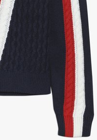 Granatowy dzianinowy sweter z rękawem, na którym biegną pionowo białe i czerwone paski w warkocz oraz teksturowane wzory warkoczowe na materiale.