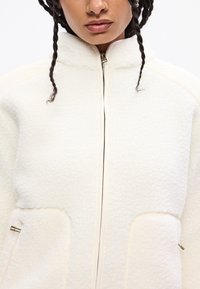 Giacca in pile bianca con colletto alto, chiusura con zip e due tasche frontali. Il tessuto ha una superficie morbida e testurizzata, accentuata da dettagli color crema.