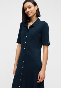 Γυναίκα που φοράει ένα φόρεμα μπλε ναυτικό με κοντά μανίκια και κουμπιά στο μπροστινό μέρος με γιακά, στέκεται μπροστά σε ένα απλό φωτεινό φόντο.