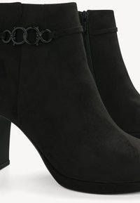 Botas de tornozelo em camurça preta com um salto chunky, apresentando um detalhe decorativo em corrente em forma de círculo e um fecho de correr lateral para fácil calçar.